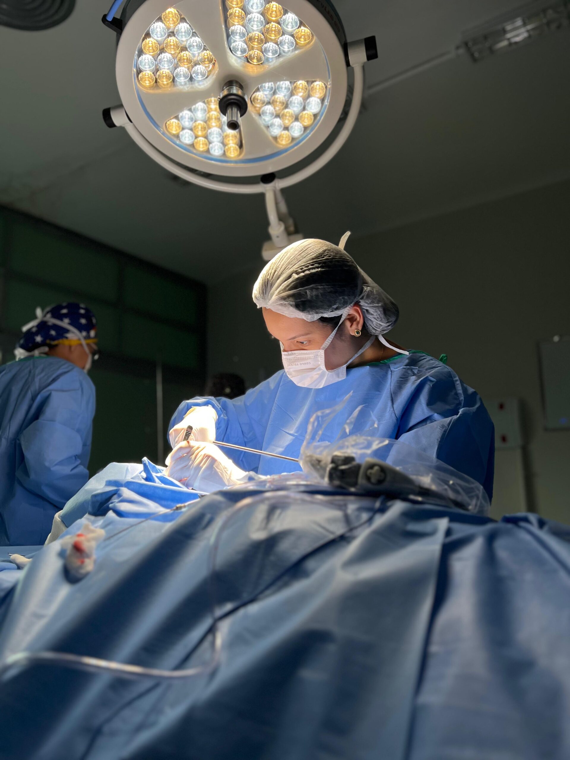 Cirurgia sendo realizada em ambiente hospitalar com profissionais usando trajes estéreis e iluminação cirúrgica.