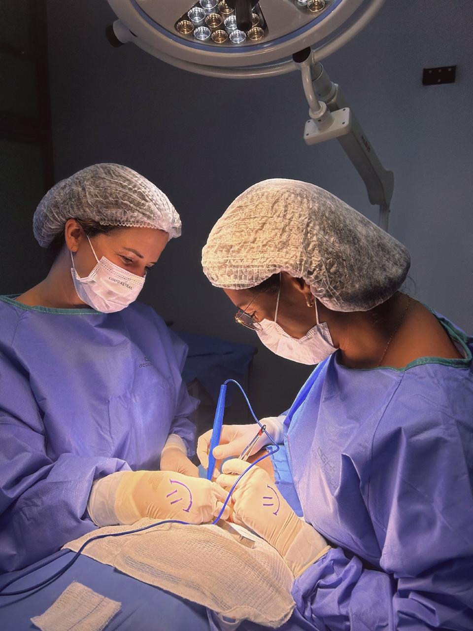 Duas profissionais de saúde realizando uma cirurgia em um ambiente hospitalar, usando trajes estéreis e focadas no procedimento.